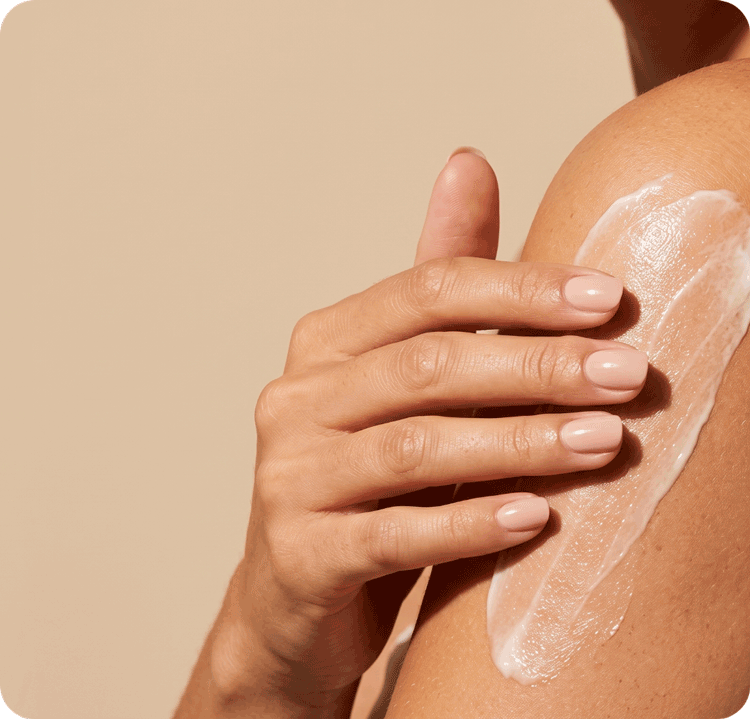 A close-up of a person's hand applying a smooth layer of white lotion or moisturizer onto their upper arm against a neutral beige background.